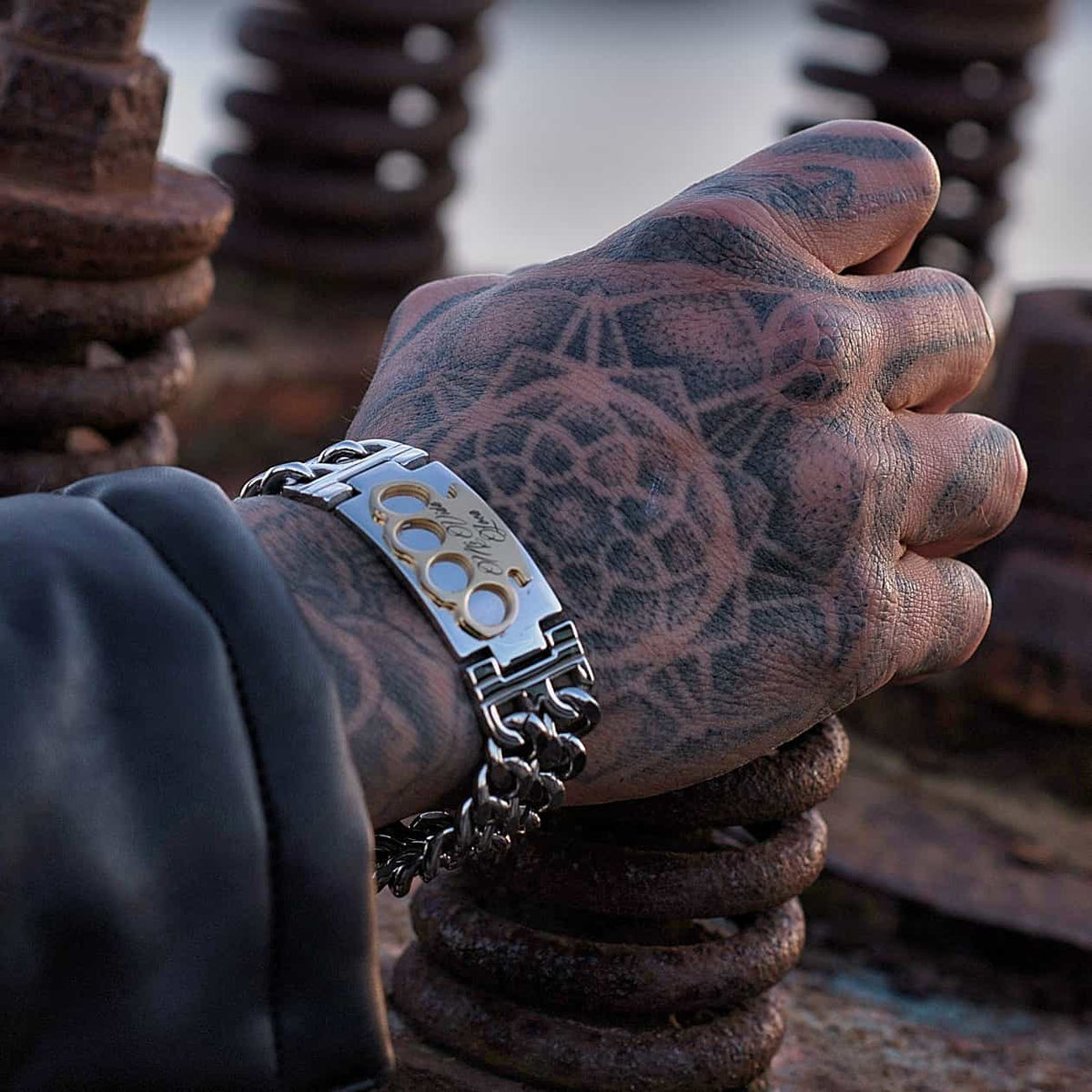 Bracciale MVL argento/oro su una mano tatuata sopra spirali di metallo arrugginito