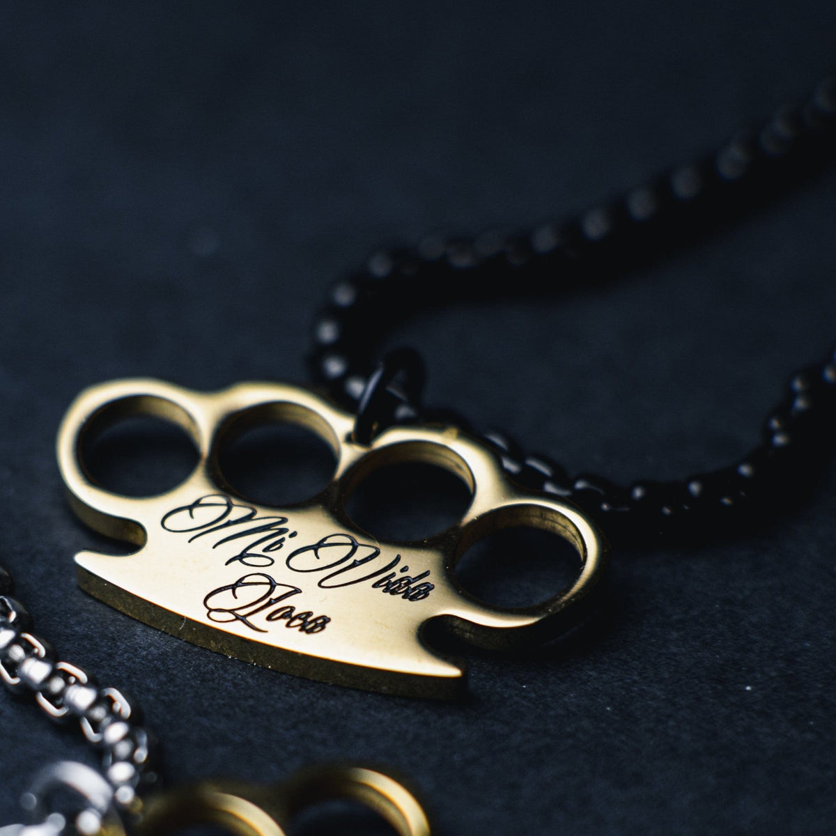 Nahaufnahme des goldenen MVL-Logo-Anhängers mit gravierter Mi Vida Loca Schrift an der schwarzen Kette